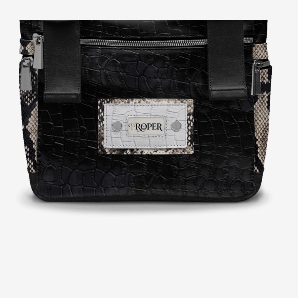 Close-up of the PROPER Signature Backpack in BLACK CROC: PROPER plate on white croc texture with snake-textured trim and silver-tone zippers